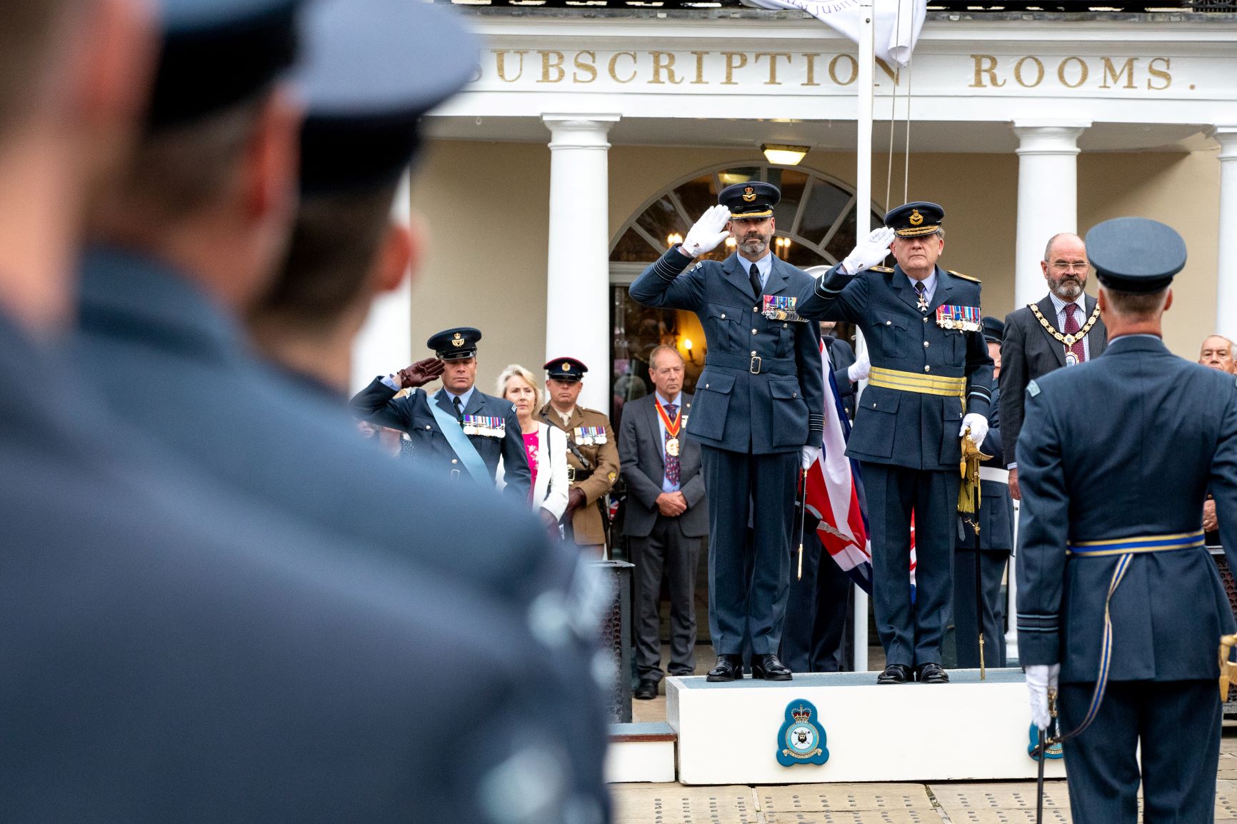 RAF Aviators salute during parade upon tiered stage.